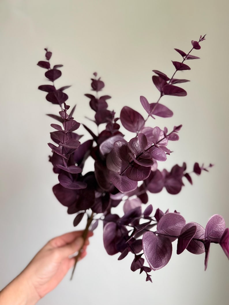 May include: A decorative stem of artificial eucalyptus featuring multiple branches with rounded, deep purple leaves. The foliage displays a matte texture and consistent dark violet coloration throughout the entire plant structure.
