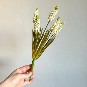 May include: A bundle of artificial flowers with white, textured blooms and green tips. The stems are green, and the leaves are long and olive-green. The flowers are held in a hand against a light gray background.