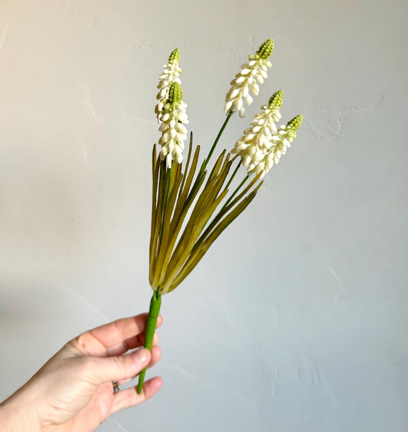 May include: A bundle of artificial flowers with white, textured blooms and green tips. The stems are green, and the leaves are long and olive-green. The flowers are held in a hand against a light gray background.