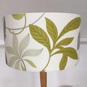 May include: A white lampshade with a green and gray leaf pattern. The lampshade is cylindrical and has a wooden base.
