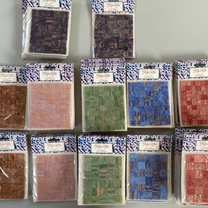 May include: Ten packages of small, colorful mosaic tiles. Each package contains a different pattern of tiles in shades of brown, pink, green, blue, and red. The packages are labeled "Tile Cafe Craft Collection".