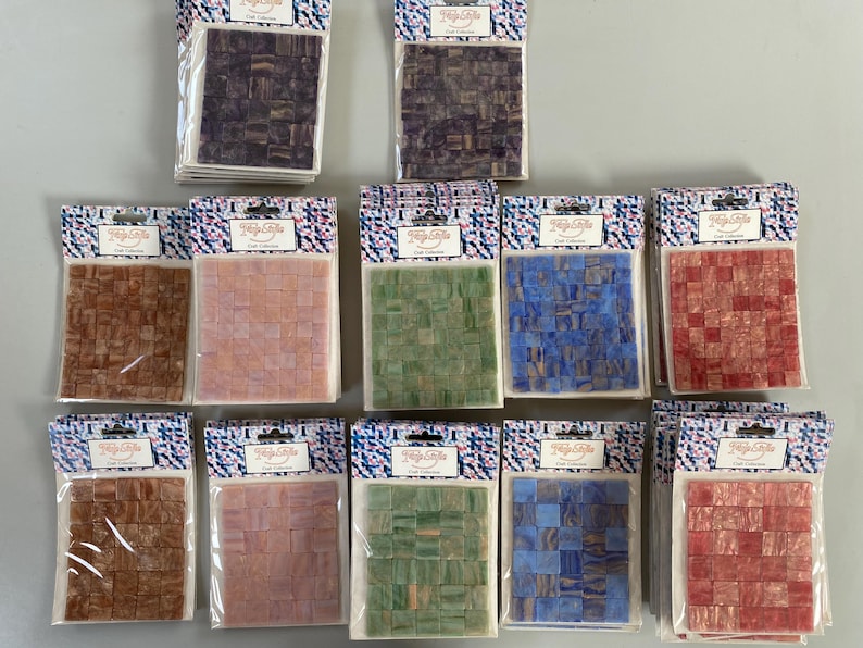 May include: Ten packages of small, colorful mosaic tiles. Each package contains a different pattern of tiles in shades of brown, pink, green, blue, and red. The packages are labeled "Tile Cafe Craft Collection".