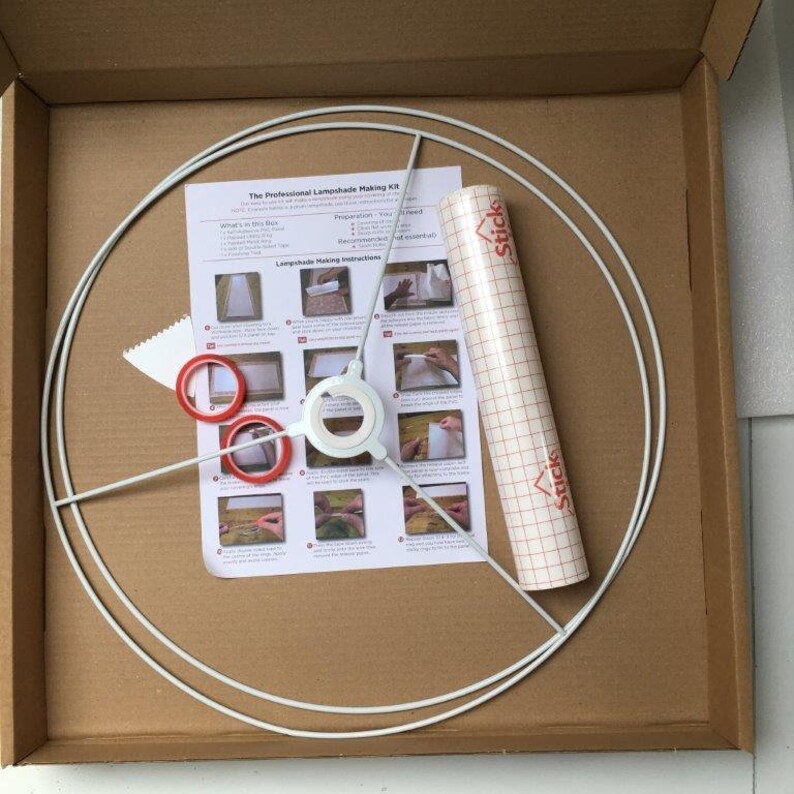 May include: A white lampshade making kit in a brown cardboard box. The kit includes a white metal lampshade frame, a roll of clear adhesive-backed plastic, a sheet of instructions, and three red rings.