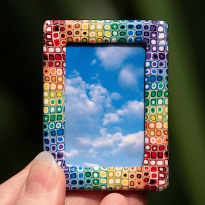 May include: A small, colorful picture frame with a rainbow pattern. The frame is holding a photo of a blue sky with white clouds.