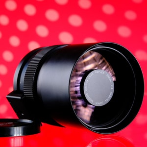Puede incluir: Un objetivo Yashica Reflex 500mm f/8 negro con una tapa de objetivo, sobre un fondo rojo con lunares blancos. El objetivo tiene un anillo de enfoque estriado y una etiqueta plateada con texto.