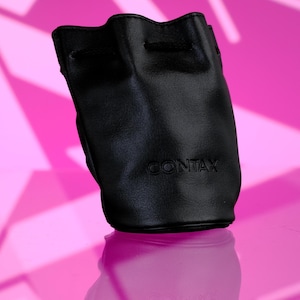 May include: Black leather Contax lens pouch with a drawstring closure. The pouch has the Contax logo embossed on the front. The background is a pink and white geometric pattern.