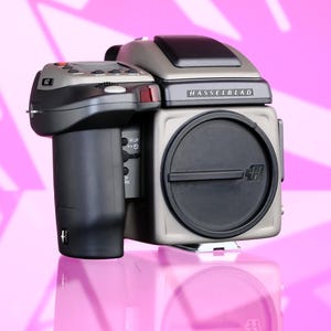 May include: A medium format Hasselblad camera with a gray body and black accents. The camera has a large lens and a top-mounted viewfinder. The brand name "Hasselblad" is visible on the top. The camera is on a reflective pink surface.