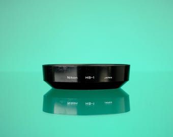 May include: Black Nikon HB-1 lens hood with the text "Nikon HB-1 JAPAN" in white. The lens hood is sitting on a reflective surface, with a teal background.