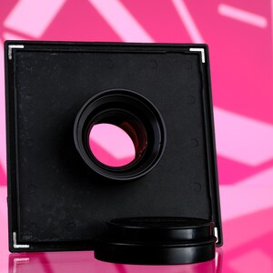 Puede incluir: Un objetivo de c&aacute;mara negro con un marco cuadrado y una abertura circular. El objetivo se encuentra sobre un fondo rosa con formas geom&eacute;tricas. Una tapa de objetivo negra se encuentra debajo.