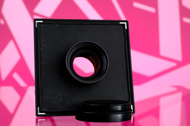 Puede incluir: Un objetivo de c&aacute;mara negro con un marco cuadrado y una abertura circular. El objetivo se encuentra sobre un fondo rosa con formas geom&eacute;tricas. Una tapa de objetivo negra se encuentra debajo.