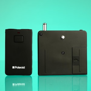 May include: Two black Polaroid camera accessories are displayed against a teal background. The item on the left is rectangular with a button and the Polaroid logo. The item on the right is a square with a silver pin and a small compartment.