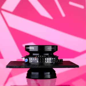 Puede incluir: Un objetivo Copal negro con la marca Rodenstock, montado sobre una base negra. La lente tiene varias marcas y ajustes num&eacute;ricos. El fondo es rosa vibrante con un patr&oacute;n geom&eacute;trico.