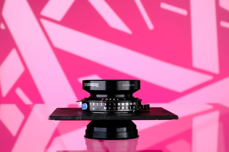 Puede incluir: Un objetivo Copal negro con la marca Rodenstock, montado sobre una base negra. La lente tiene varias marcas y ajustes num&eacute;ricos. El fondo es rosa vibrante con un patr&oacute;n geom&eacute;trico.