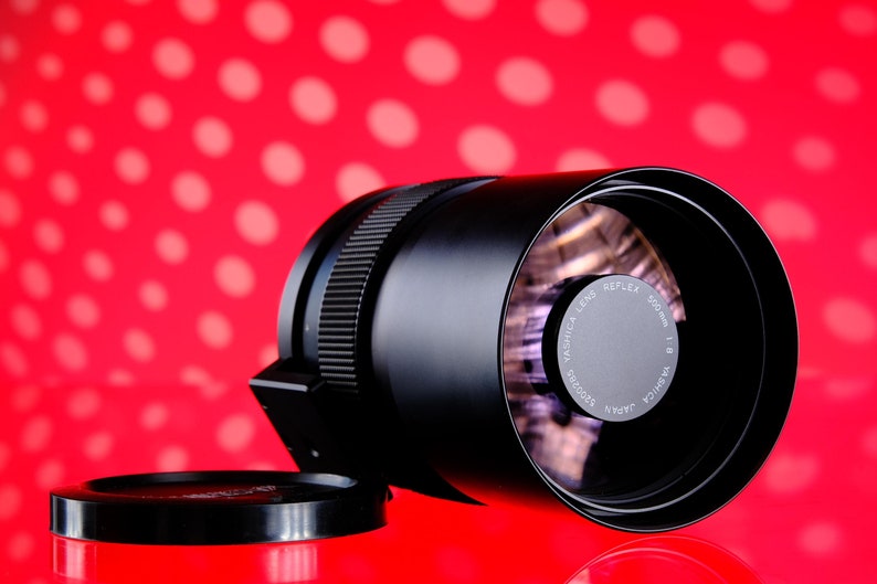 Puede incluir: Un objetivo Yashica Reflex 500mm f/8 negro con una tapa de objetivo, sobre un fondo rojo con lunares blancos. El objetivo tiene un anillo de enfoque estriado y una etiqueta plateada con texto.