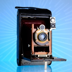 May include: Vintage wooden and leather camera with a lens and flash attachment. The camera has a brown wooden body, a black leather case, and a silver metal hinge. The camera is open, revealing the bellows and lens.