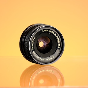 May include: A Konica Hexanon AR 28mm F3.5 camera lens, made in Japan, with a black and silver finish. The lens is reflected on a glossy orange surface. The lens has a ribbed focus ring and a clear glass element.