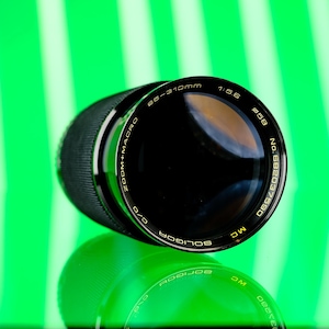 Puede incluir: Un objetivo de cámara zoom macro Soligor negro con el texto "80-310mm 1:5.6" y "MC" impreso en el objetivo. El objetivo está sobre un fondo verde brillante con un reflejo del objetivo debajo.