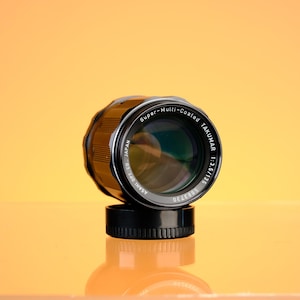 May include: A black and silver Super-Multi-Coated Takumar 1:3.5/135 camera lens. The lens is mounted on a black base, with the lens barrel featuring a ribbed design. The text "ASAHI OPT. CO. JAPAN" is visible on the lens.