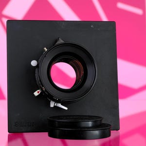 Puede incluir: Un objetivo de c&aacute;mara negro con una base cuadrada y dos tapas de objetivo. El objetivo tiene una abertura circular con un reflejo rosa. La base tiene la palabra "Sinar" impresa. El fondo es rosa brillante con formas geom&eacute;tricas.