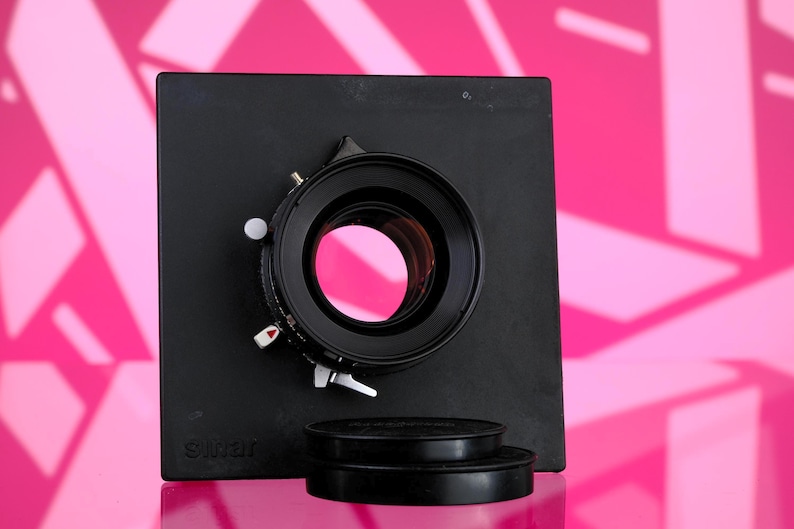 Puede incluir: Un objetivo de c&aacute;mara negro con una base cuadrada y dos tapas de objetivo. El objetivo tiene una abertura circular con un reflejo rosa. La base tiene la palabra "Sinar" impresa. El fondo es rosa brillante con formas geom&eacute;tricas.
