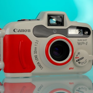 May include: A Canon Sure Shot WP-1 camera, with a white body and red accents, and a red wrist strap. The camera has a 32mm lens. The word "Canon" is printed on the front.