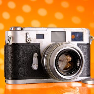 Può includere: Una macchina fotografica a pellicola da 35 mm argentata e nera con un copriobiettivo. La macchina fotografica ha un rivestimento in pelle nera e un obiettivo argentato con un anello nero attorno. La macchina fotografica è appoggiata su uno sfondo arancione brillante.