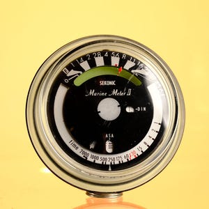 Puede incluir: Un exposímetro redondo Sekonic Marine Meter II con esfera negra, escalas numéricas blancas y un indicador de arco verde. El dispositivo presenta texto en blanco que incluye 'SEKONIC', 'Marine Meter II', 'ASA' y 'time' con varios números.