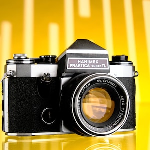 May include: A black and silver 35mm film camera with a silver lens. The camera has the text "HANIMEX PRAKTICA super TL" on the top and "AUTO YASHINON DX - 1:1.8 50mm No. 54106411 JAPAN" on the lens.