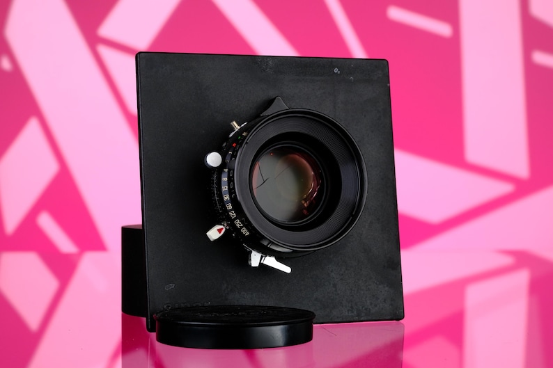 Puede incluir: Un objetivo de c&aacute;mara negro con un dial de control plateado y blanco y una tapa de objetivo negra. El objetivo est&aacute; montado en una placa cuadrada negra. El fondo es rosa brillante con un patr&oacute;n geom&eacute;trico.