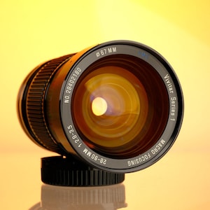 May include: A Vivitar Series 1 macro focusing camera lens with a black body and a 67mm diameter. The lens has a 28-90mm 1:2.8-3.5 focal length and is set against a gradient yellow and orange background.
