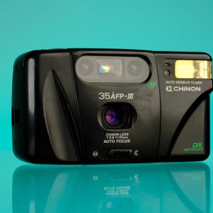 Puede incluir: Cámara Chinon 35AFP-III negra con flash incorporado. La cámara tiene una lente con el texto "CHINON LENS 1:3.9 f=35mm AUTO FOCUS". La cámara está sobre una superficie reflectante con fondo turquesa.
