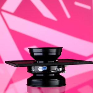 Puede incluir: Un objetivo de c&aacute;mara negro con una base cuadrada y una parte superior cil&iacute;ndrica. El objetivo se encuentra sobre un fondo rosa vibrante con formas geom&eacute;tricas blancas. El objetivo tiene un dial con texto y una pegatina.