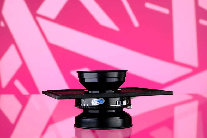 Puede incluir: Un objetivo de c&aacute;mara negro con una base cuadrada y una parte superior cil&iacute;ndrica. El objetivo se encuentra sobre un fondo rosa vibrante con formas geom&eacute;tricas blancas. El objetivo tiene un dial con texto y una pegatina.