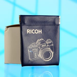 Puede incluir: Una funda de cuero azul marino con la palabra "RICOH" y una ilustración de cámara dorada. La funda tiene un cierre metálico dorado en la parte superior. Una caja beige es parcialmente visible en el fondo.