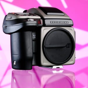 Könnte beinhalten: Eine Mittelformatkamera von Hasselblad mit schwarzem Objektiv und silbernem und schwarzem Gehäuse. Auf der Kamera steht das Wort "Hasselblad". Der Hintergrund ist ein rosa-weißes geometrisches Muster.