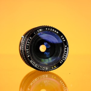 Puede incluir: Un objetivo de cámara Soligor wide-auto con una distancia focal de 28 mm y una apertura de f/2.8, sobre un fondo naranja vibrante. El objetivo es negro con detalles plateados y presenta texto visible, incluido el número de serie y los detalles del modelo. El objetivo se refleja en la superficie.