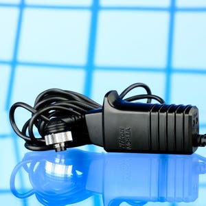 May include: A black Nikon MD-12A camera battery charger with a coiled cord and a silver-colored connector. The charger is on a reflective surface with a blue background.