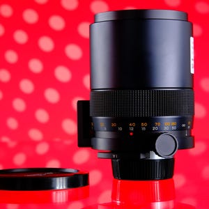 Puede incluir: Un objetivo de c&aacute;mara negro con un agarre texturizado y marcas de medici&oacute;n. El objetivo est&aacute; sobre un fondo rojo con lunares blancos, y una tapa de objetivo es visible en primer plano.
