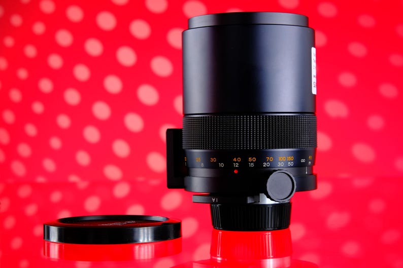 Puede incluir: Un objetivo de c&aacute;mara negro con un agarre texturizado y marcas de medici&oacute;n. El objetivo est&aacute; sobre un fondo rojo con lunares blancos, y una tapa de objetivo es visible en primer plano.