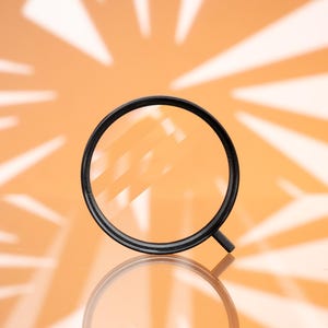 May include: A black magnifying glass with a circular lens and a short handle, set against an orange background with a radial pattern of light and shadow. The glass is reflected on the surface below.