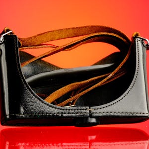 May include: Black leather camera case with brown leather straps. The case has a glossy finish and a small, black knob on the bottom. The straps are looped inside the case.