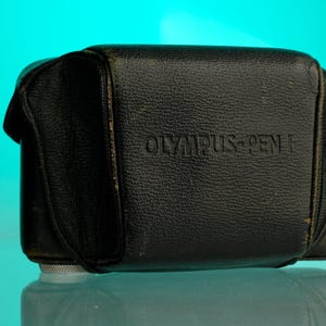 Puede incluir: Funda de cámara de cuero negro con el texto en relieve "OLYMPUS-PEN F". La funda es rectangular con bordes redondeados y una superficie texturizada. La funda tiene una base de metal plateado.