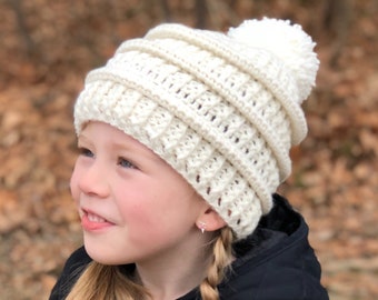 May include: A young girl wearing a white crocheted beanie with a white pom pom on top. She is wearing a black quilted jacket and has her hair in two braids.