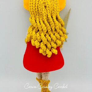 May include: A crocheted doll with long yellow hair, a red cape, and brown boots. The doll is holding a sword in its right hand. The doll is standing on a white background. The text "Crown Country Crochet" is visible at the bottom of the image.