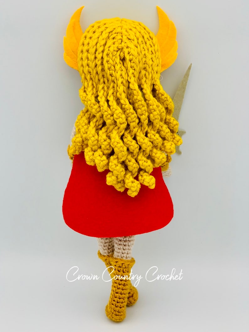 May include: A crocheted doll with long yellow hair, a red cape, and brown boots. The doll is holding a sword in its right hand. The doll is standing on a white background. The text "Crown Country Crochet" is visible at the bottom of the image.