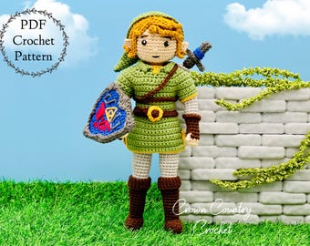 May include: Crocheted figure of a green-clad character holding a shield with a red, blue, and white Triforce symbol. The character has brown hair, a green tunic, and brown boots. The figure is standing in front of a white brick wall with green vines.