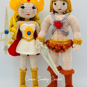May include: Crocheted figures of He-Man and She-Ra, both with blonde hair and wearing their iconic outfits. He-Man is holding a sword and She-Ra is holding a sword and shield. The figures are standing on a white background. Crown Country Crochet.