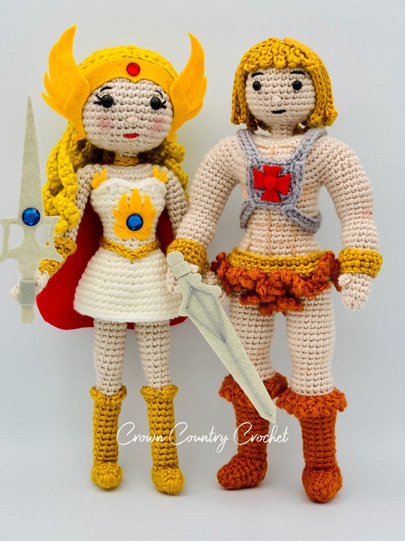 May include: Crocheted figures of He-Man and She-Ra, both with blonde hair and wearing their iconic outfits. He-Man is holding a sword and She-Ra is holding a sword and shield. The figures are standing on a white background. Crown Country Crochet.