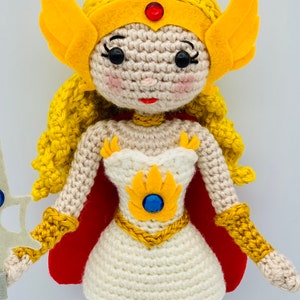 May include: Crocheted doll with yellow hair, a white dress, a red cape, and a gold crown. The doll is holding a sword.