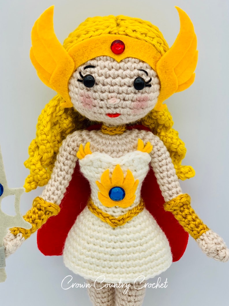 May include: Crocheted doll with yellow hair, a white dress, a red cape, and a gold crown. The doll is holding a sword.